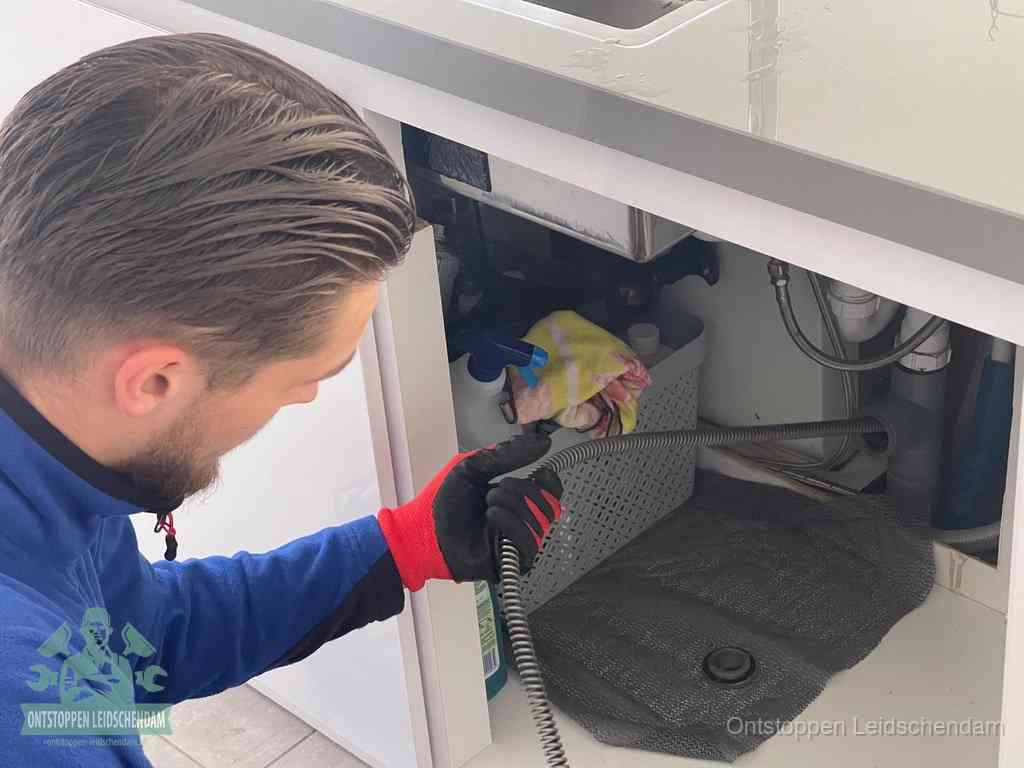 Een monteur voert een flexibele ontstoppingsveer in onder een keukenkastje om een verstopping voorbij de sifon te bereiken in Leidschendam