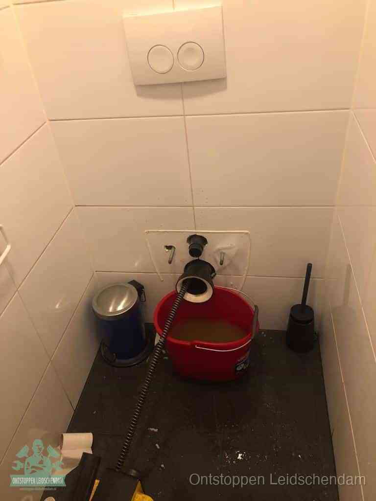 Een ontstopte wand-wc met verwijderde toiletpot waar een ontstoppingsveer in de wanduitgang verdwijnt, het achterhalen waar de verstopping zit in Leidschendam