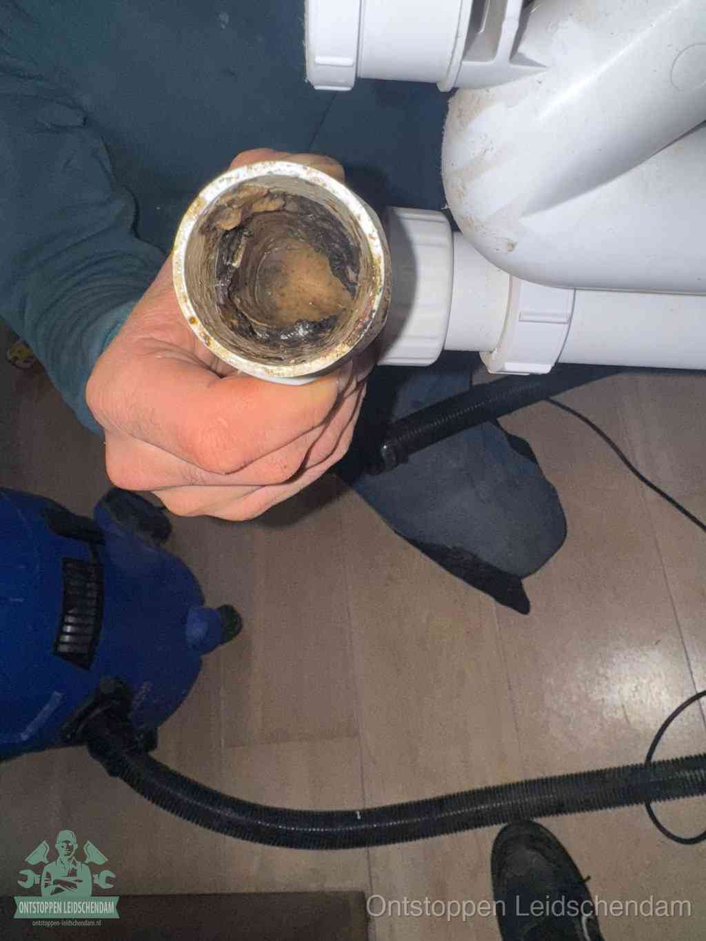 Een hand houdt een losgekoppeld PVC-afvoerbuisstuk vast dat van binnen volledig is dichtgeslibd met vet, vuil en aanslag, de meest voorkomende oorzaak van een verstopte afvoer in Leidschendam