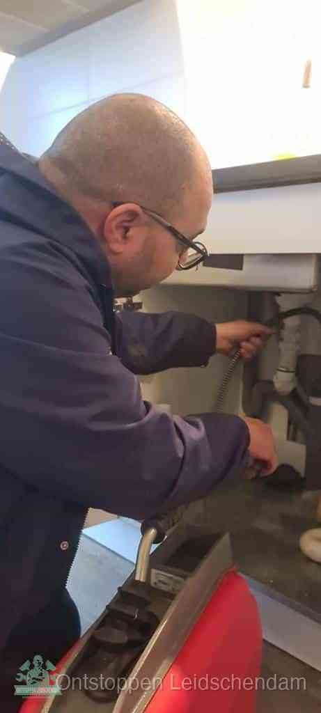 Een monteur buigt zich onder een wastafel en voert een ontstoppingsveer in om de bron van rioollucht uit de afvoer in Leidschendam op te sporen