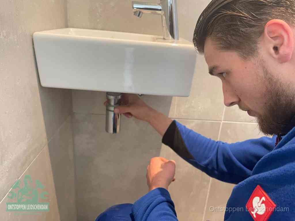 Een monteur draait met de hand de zwanenhalssifon onder een fonteinwasbak in de toiletruimte los om de bron van rioollucht in Leidschendam te vinden