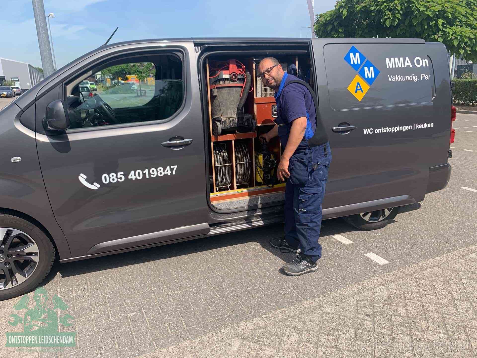 Een monteur bij de open schuifdeur van zijn bedrijfsbus op een parkeerplaats, klaar om de oorzaak van terugkerende afvoerverstoppingen aan te pakken in Leidschendam