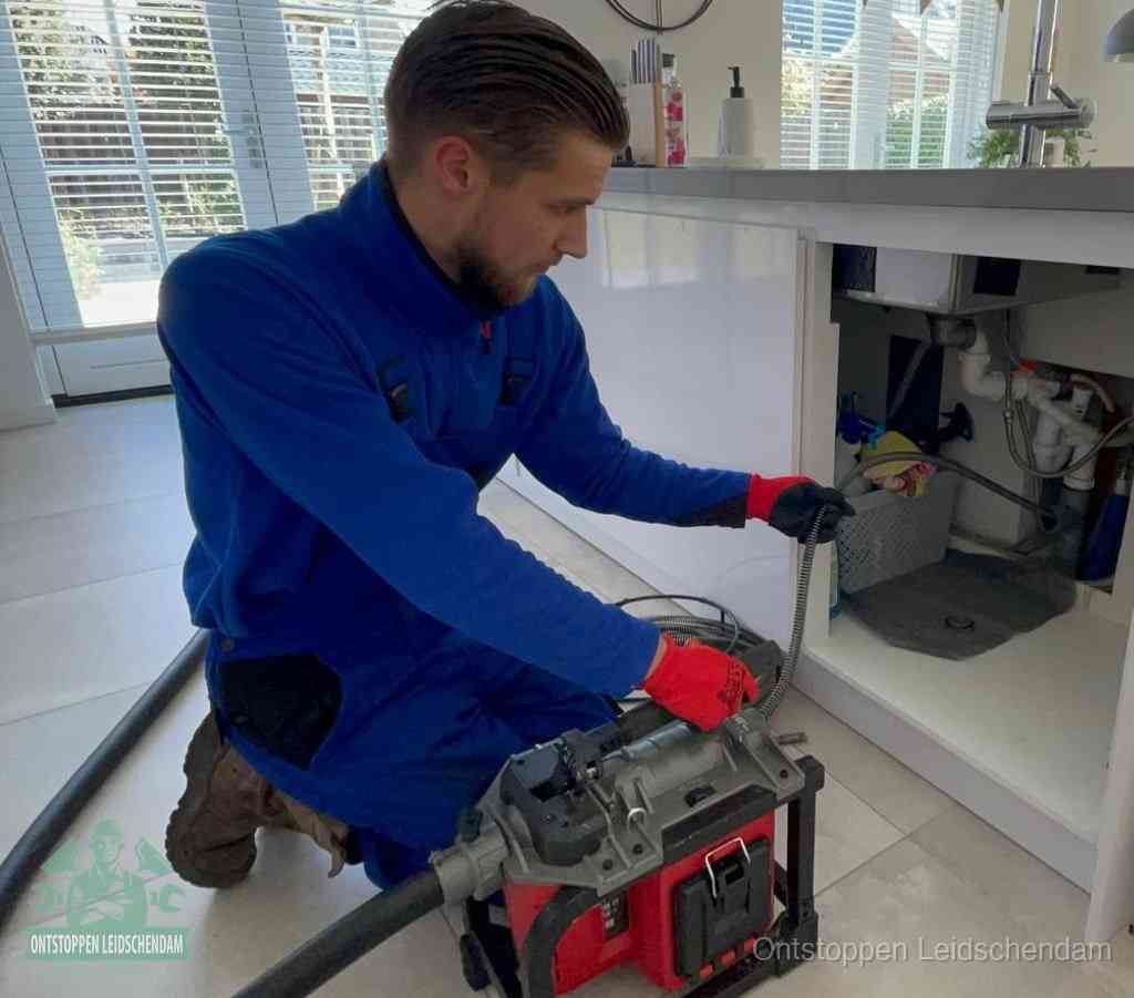 Een monteur met rode handschoenen knielt in een keuken en voert een elektrische ontstoppingsveer in tijdens het professioneel afvoer ontstoppen in Leidschendam