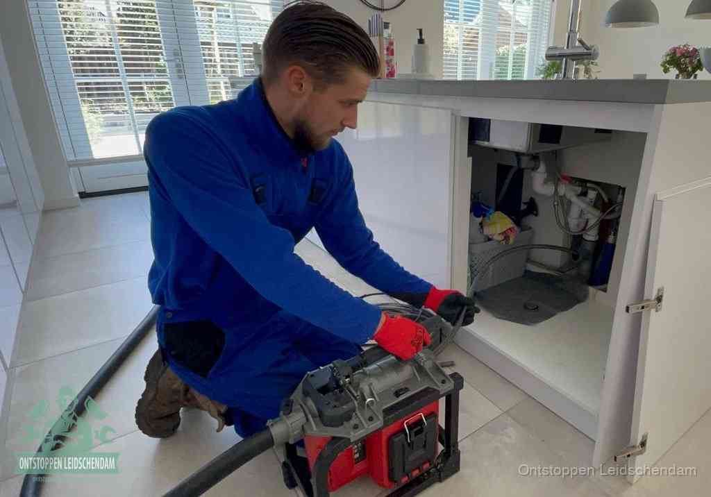 Een monteur voert een professionele elektrische ontstoppingsmachine in de afvoer onder de spoelbak tijdens het gootsteen ontstoppen in Leidschendam